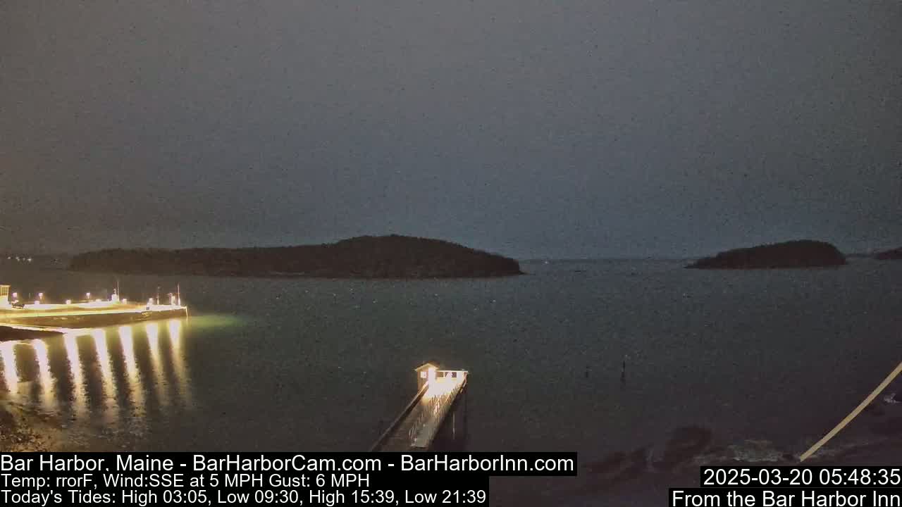A dark, nighttime scene shows a lit-up pier extending into calm water, with islands visible in the distance under an overcast sky.