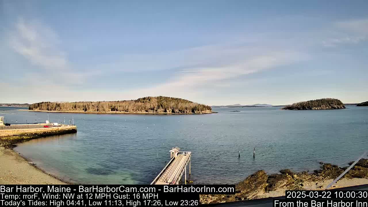 A calm, sunny day shows a long pier extending into a body of water, with several islands visible in the distance under a mostly clear sky.