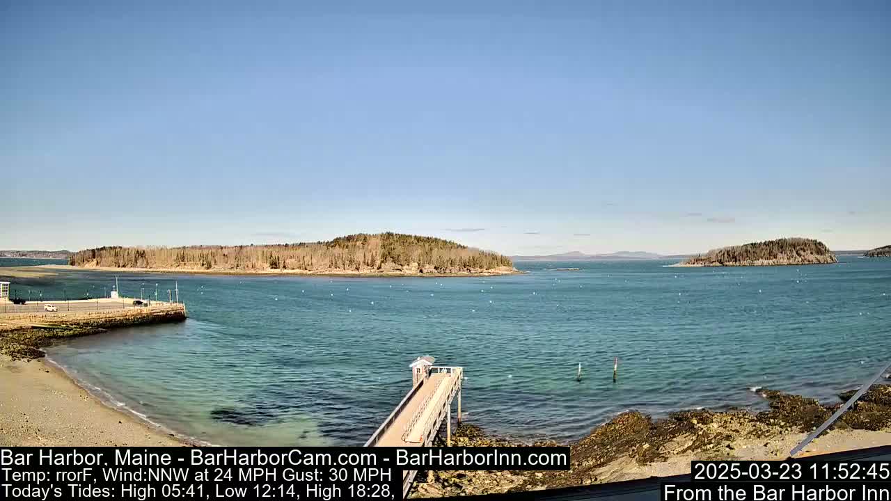 A partly sunny day shows a tranquil harbor scene with a long pier extending from a rocky shore into calm, clear water,  two islands in the distance, and a small sandy beach.