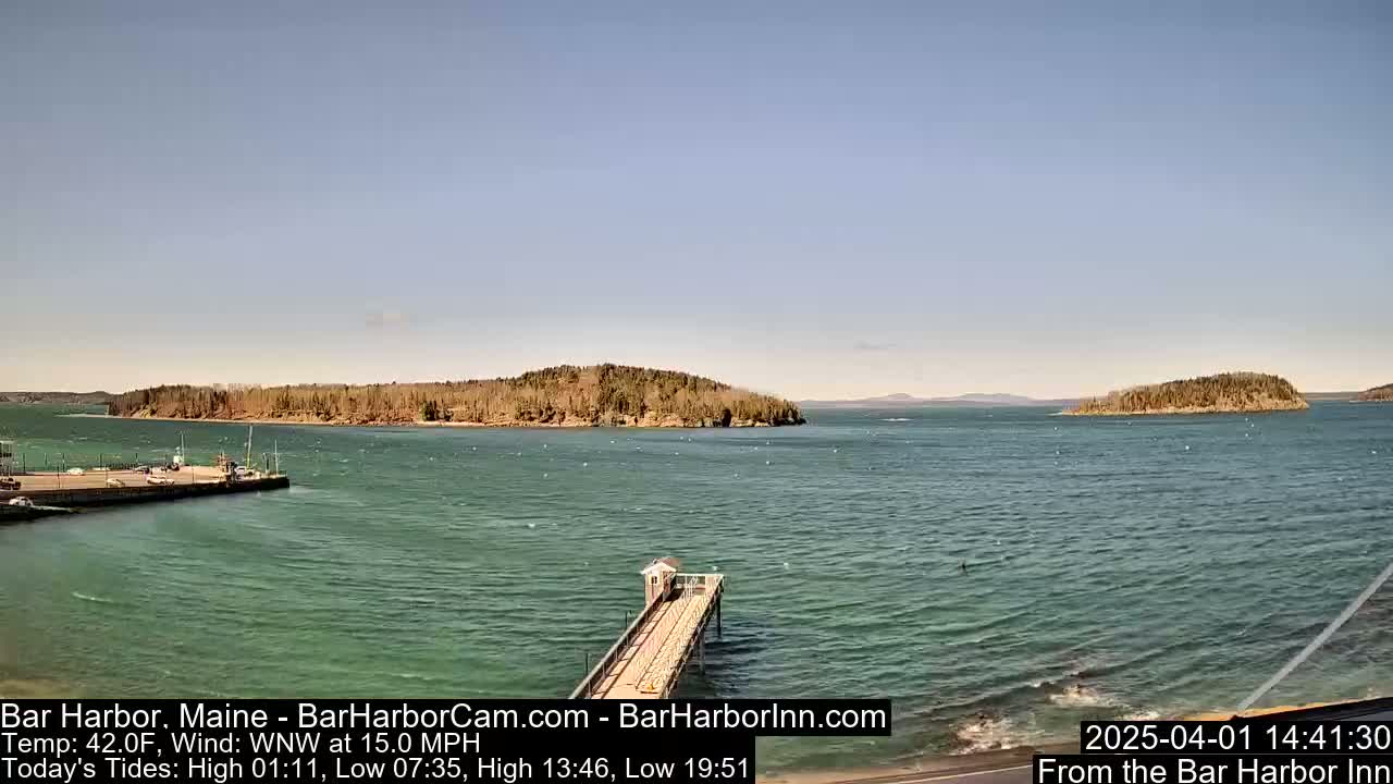 A long pier extends into a calm, teal-green body of water, with several small, tree-covered islands visible in the distance under a clear, bright sky.