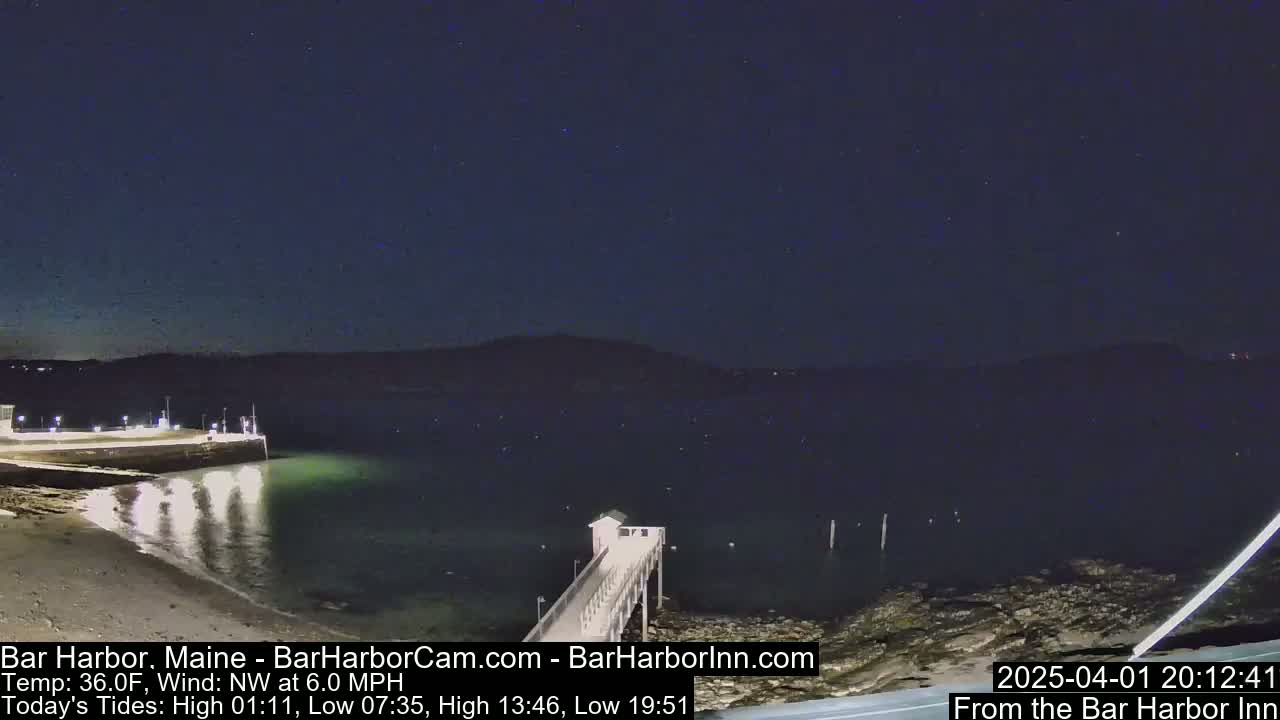 A nighttime view of a calm body of water with a pier extending from a lit shoreline under a clear, dark sky.