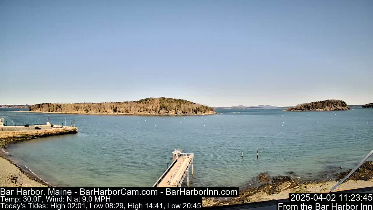 A calm, sunny day shows a harbor with a long pier extending into the water, small islands in the distance, and a shoreline with light-colored sand.