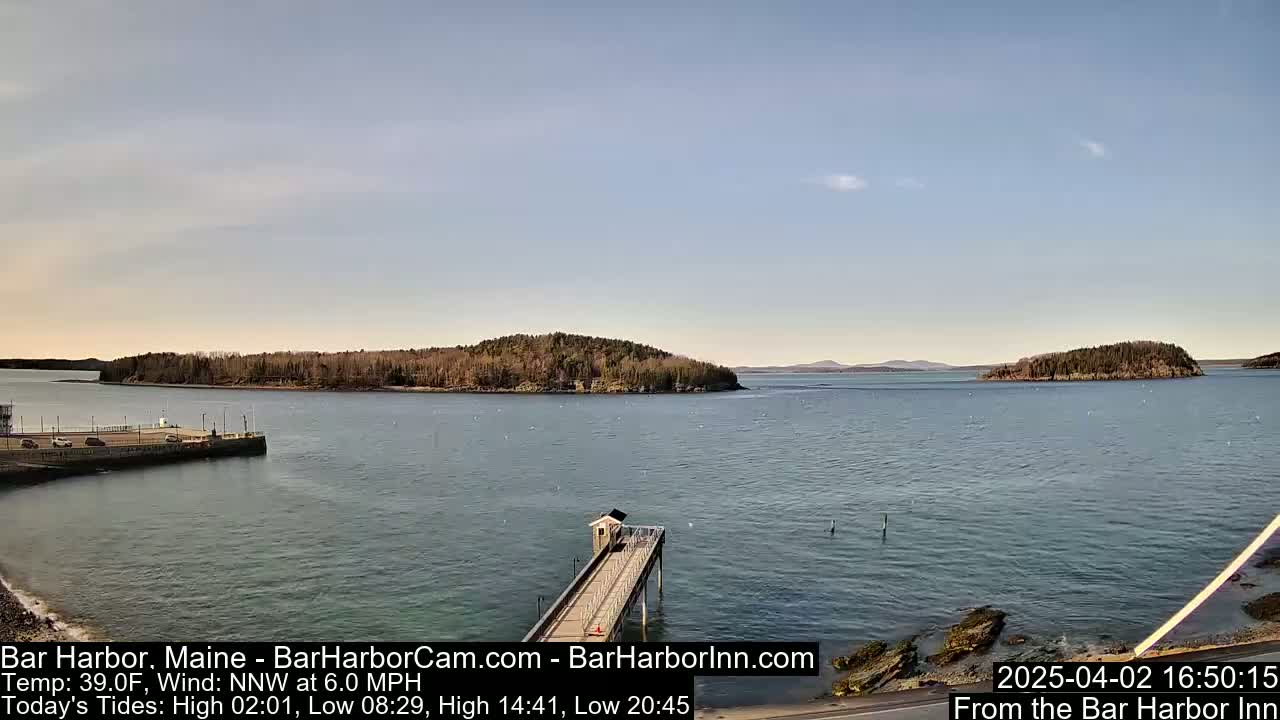 A calm body of water with a long pier extending from a shoreline, featuring islands and a distant mountain range under a clear sky.