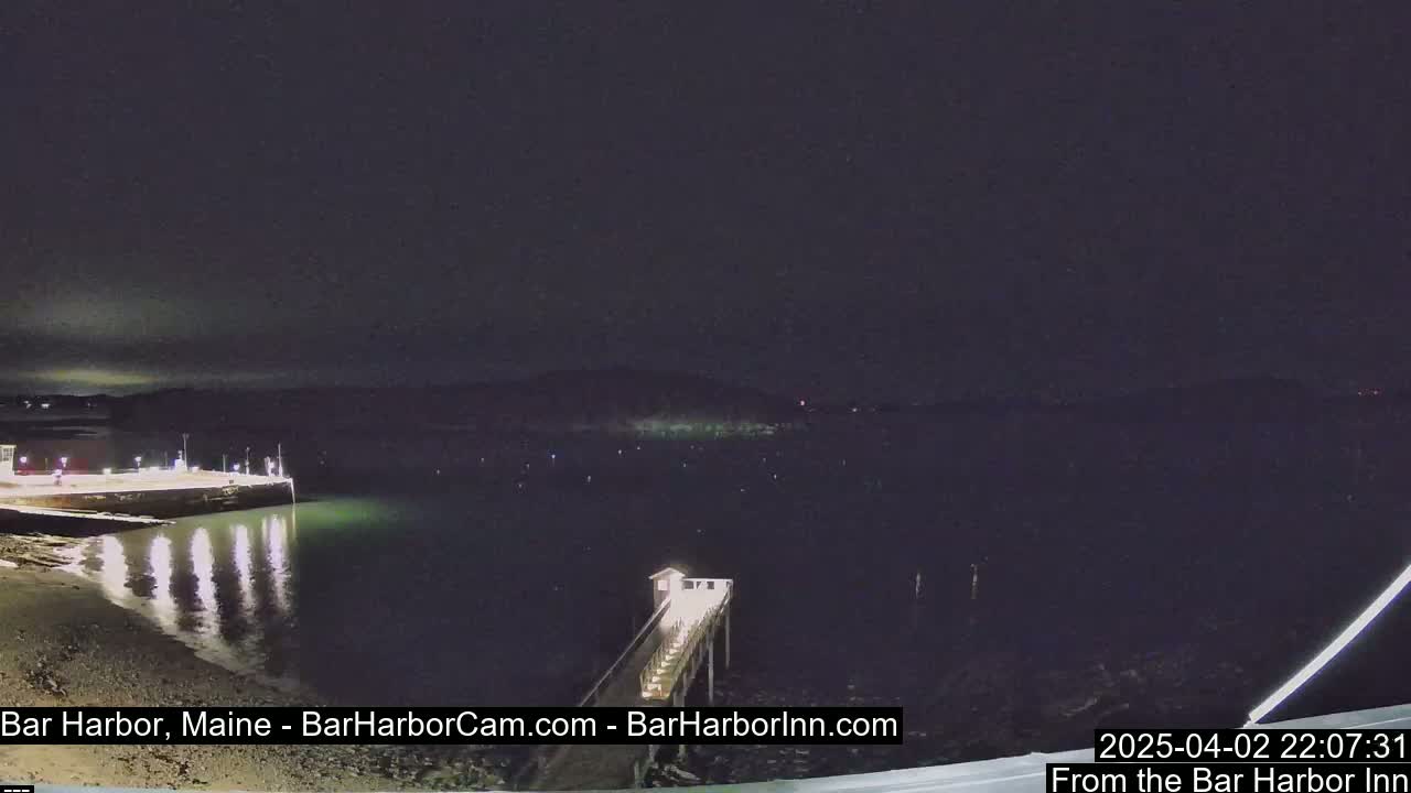 A nighttime view of a calm harbor with a pier extending from a lit structure, under a dark, cloudy sky.
