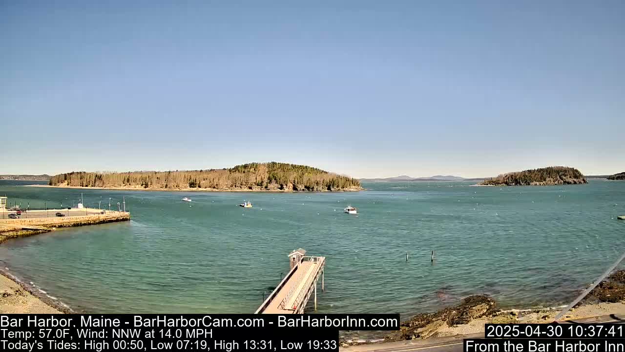 A calm, sunny day shows a harbor with a long pier extending into teal water, several small boats, and tree-covered islands in the distance.