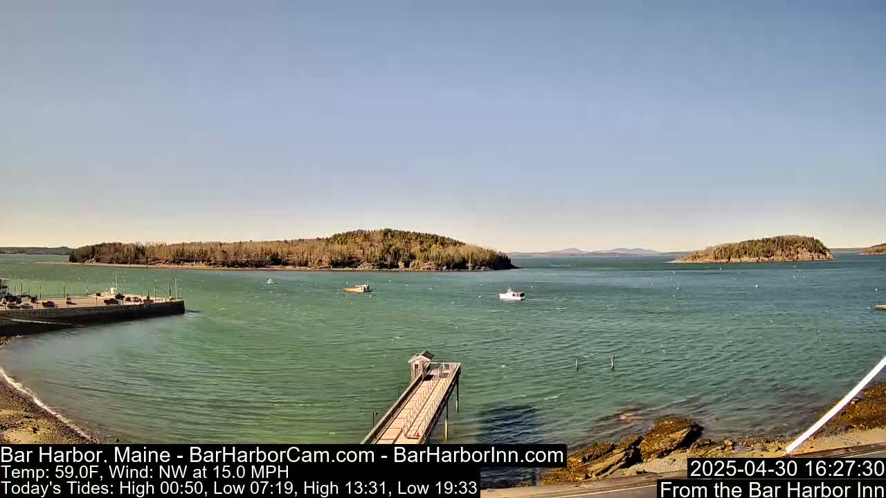 A calm, sunny day shows a harbor with a long pier, several boats, and tree-covered islands in the distance, with a northwest wind at 15 mph.