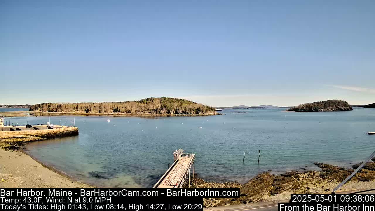 A tranquil harbor scene on a sunny day shows a calm body of water, a small sandy beach, a long pier extending into the water, and tree-covered islands in the distance.