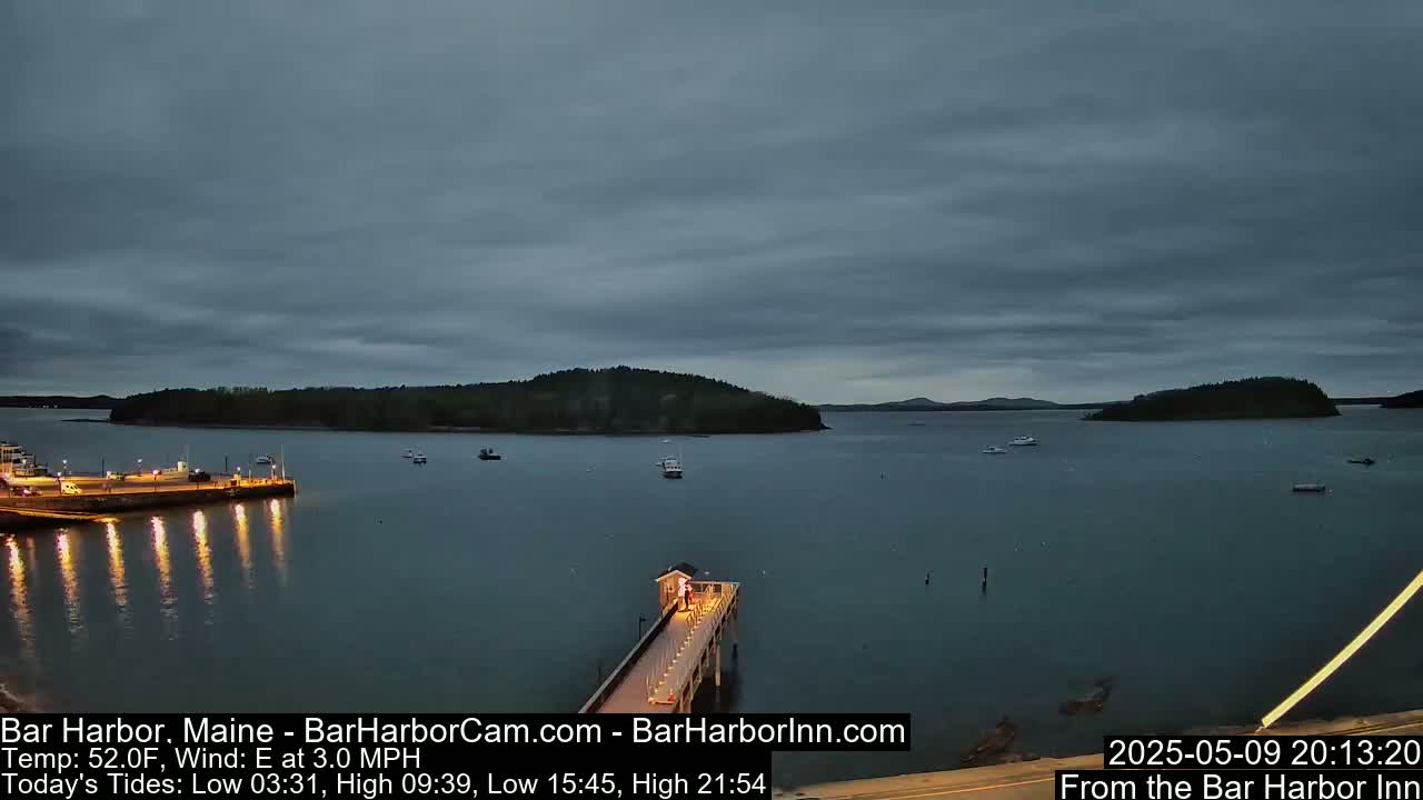A calm, twilight scene over a body of water shows a long pier extending from a shoreline with a few boats scattered across the water and islands in the distance under a cloudy sky.