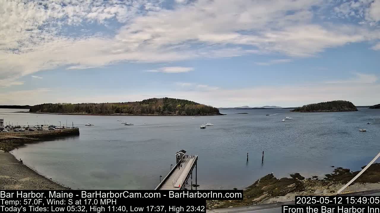 A partly cloudy sky over a calm bay with several boats and a long pier extending from a rocky shoreline, with wooded islands in the distance.