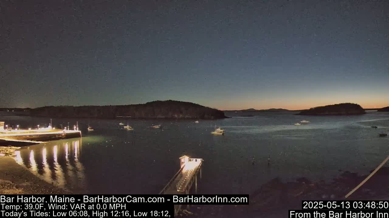 A calm, dark body of water at twilight is dotted with several boats near a lit pier and shoreline, with islands in the background.