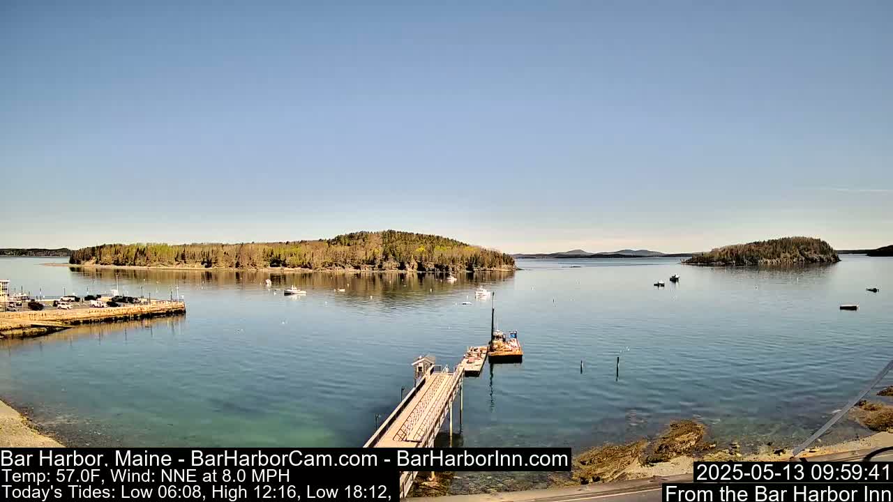 A calm harbor with a long pier, several boats, and tree-covered islands under a clear blue sky.