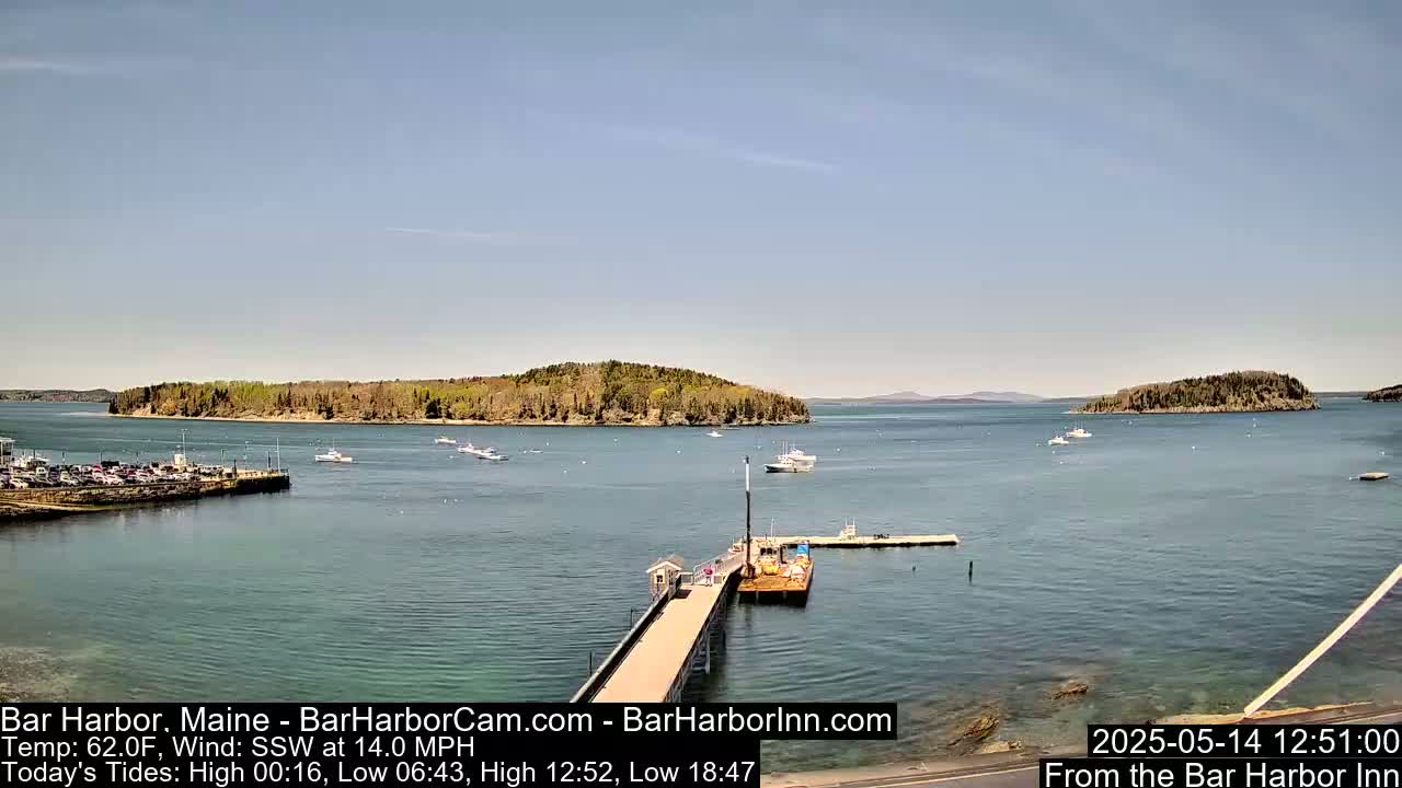 A calm harbor scene on a sunny day shows a pier extending into the water, several boats, and tree-covered islands in the distance.