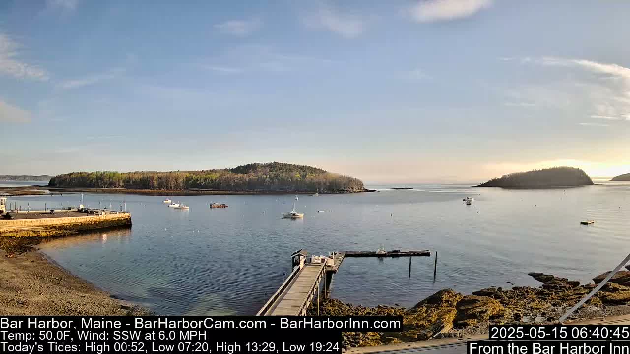 A calm bay with several small boats, a long wooden pier, and a small island in the background under a mostly clear sky.