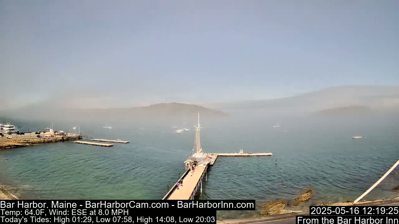 A hazy, calm harbor scene shows a long pier extending from a rocky shoreline into the water, with several boats and a sailboat docked nearby.
