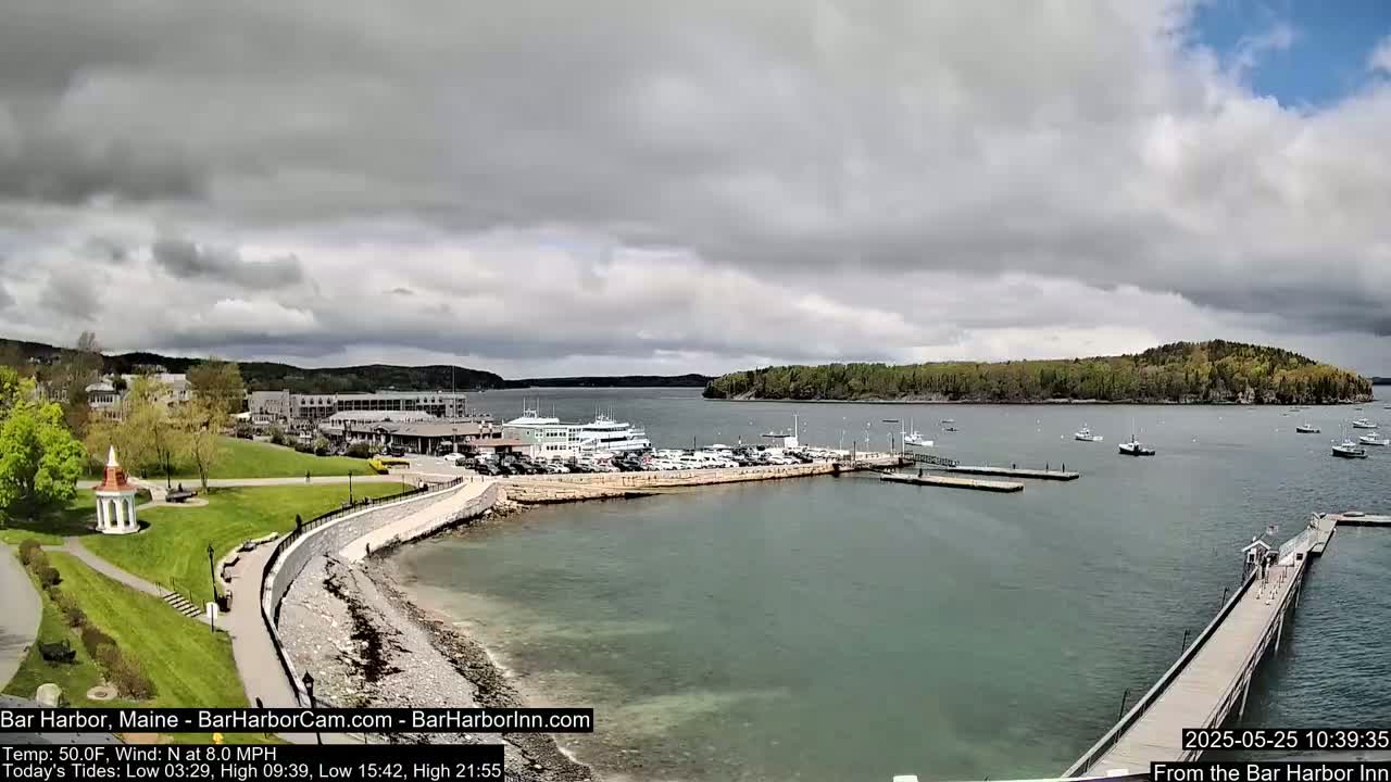 A harbor scene under partly cloudy skies shows a waterfront town with a marina filled with boats and a long pier extending into the water.