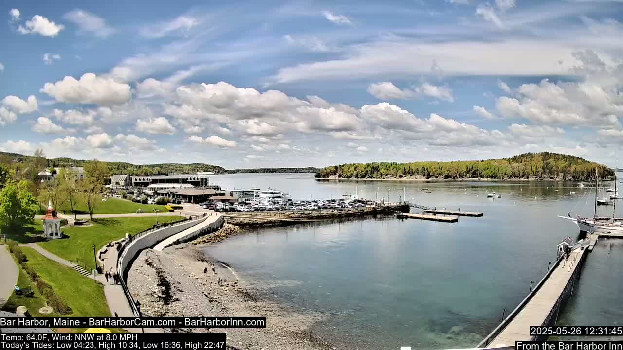 A partly cloudy day overlooks a harbor with boats, a parking lot, and a shoreline path leading to a grassy area.