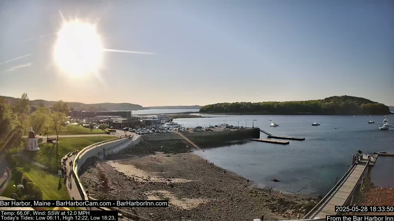 A sunny day overlooking a harbor with boats docked, a rocky shoreline, and green hills in the background.