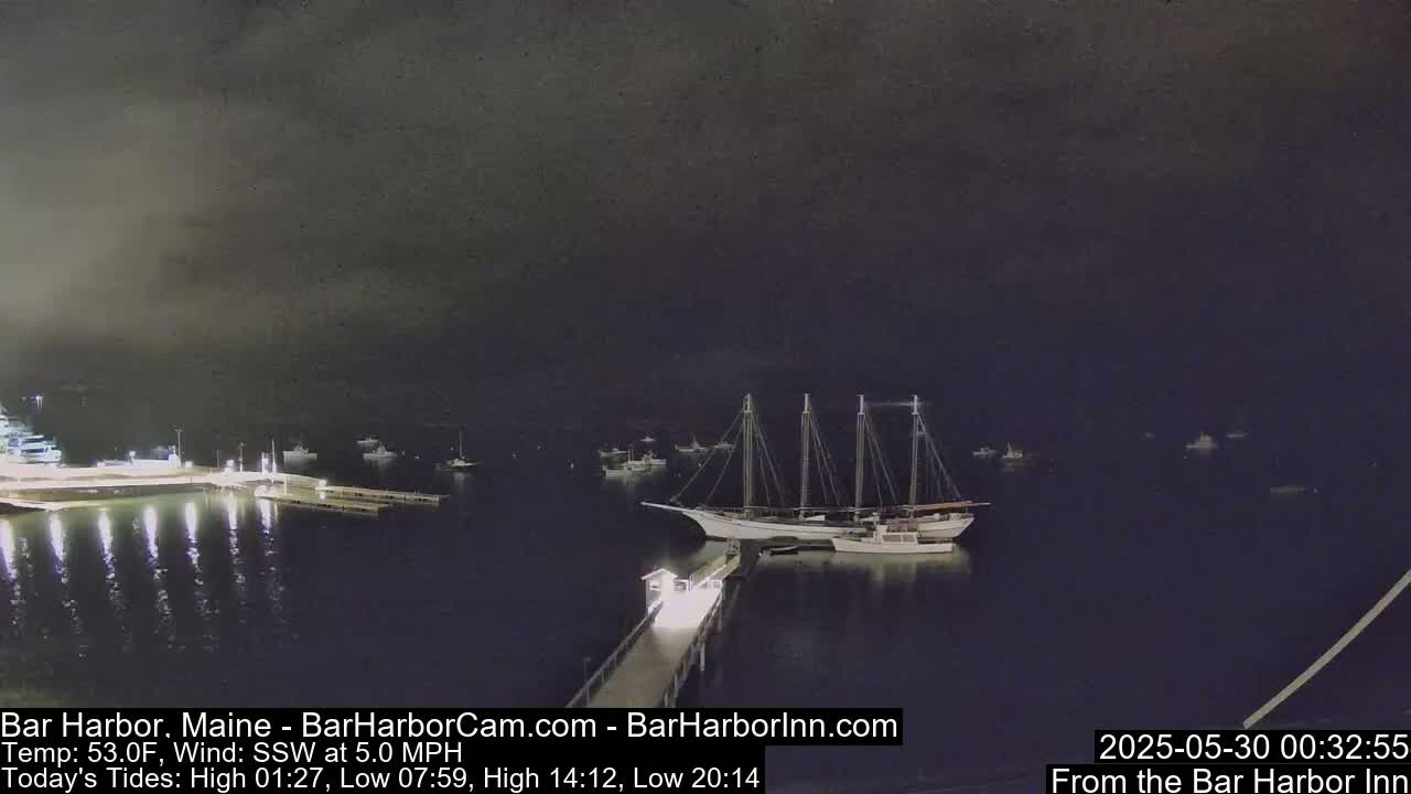 A large sailboat is moored at a dock at night, under a dark sky, with other boats visible in the harbor.