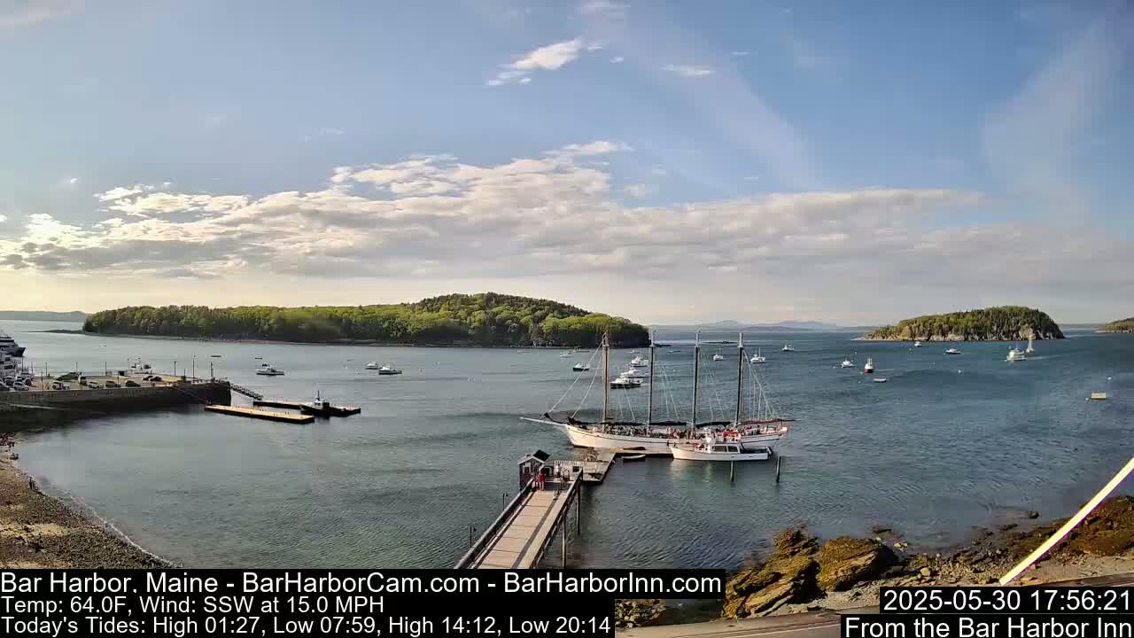 A partly cloudy day shows a harbor with numerous boats, a large sailing ship docked at a pier, and several small islands in the distance.