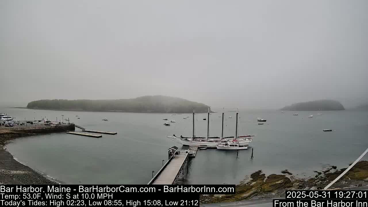 A foggy harbor scene shows a large sailboat docked at a pier, smaller boats scattered across the water, and a small island shrouded in mist in the background.