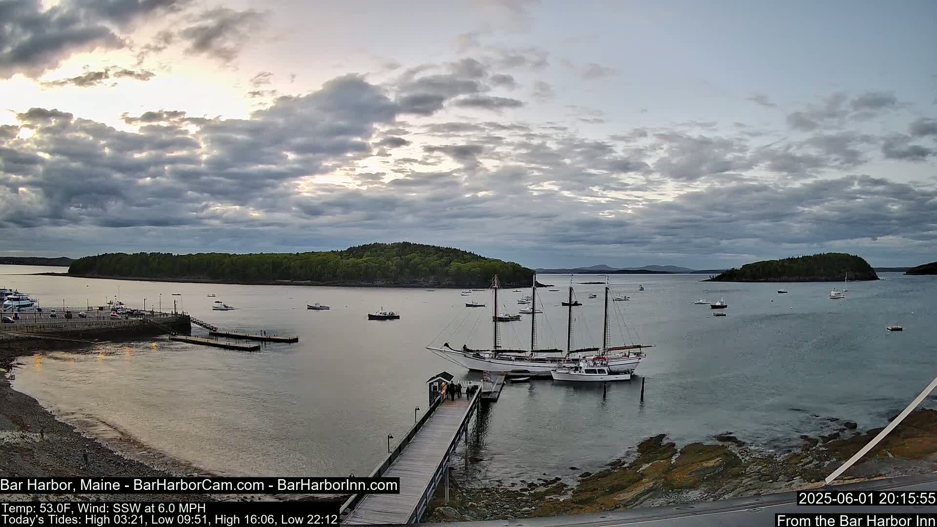 A large sailing ship is docked at a pier in a calm harbor under a partly cloudy sky, with several smaller boats and a small island visible in the distance.
