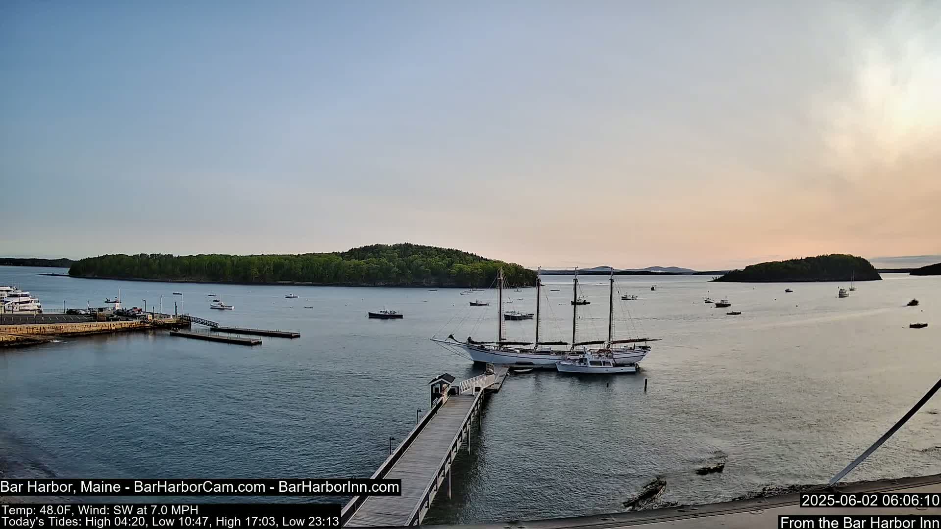 A calm harbor at sunset with several boats, including a large sailing vessel, moored at a dock, and a small island in the background.