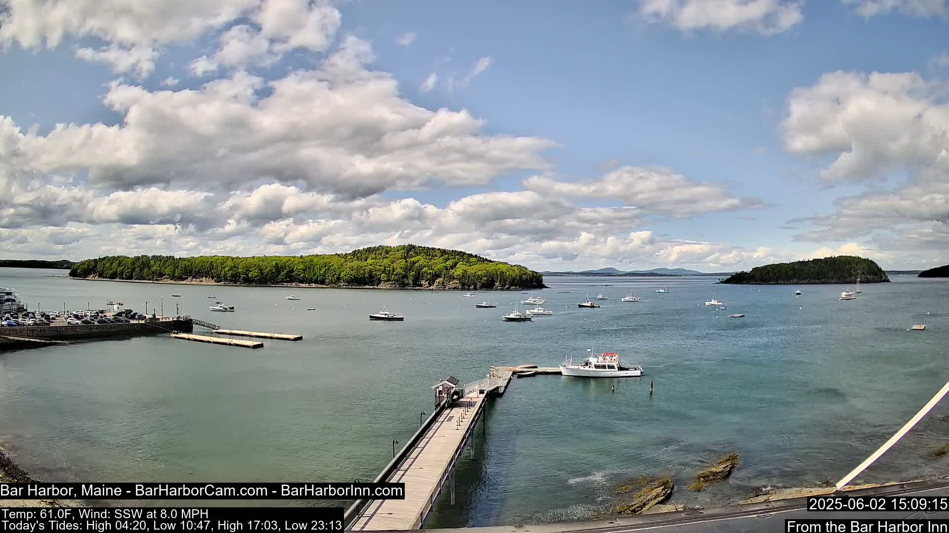 A partly cloudy day shows a harbor with numerous boats, a long dock extending into the water, and a wooded island in the distance.