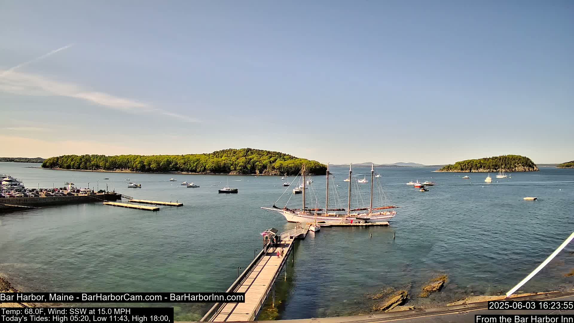A large sailing ship is docked at a pier in a calm harbor, surrounded by smaller boats and islands under a clear blue sky.
