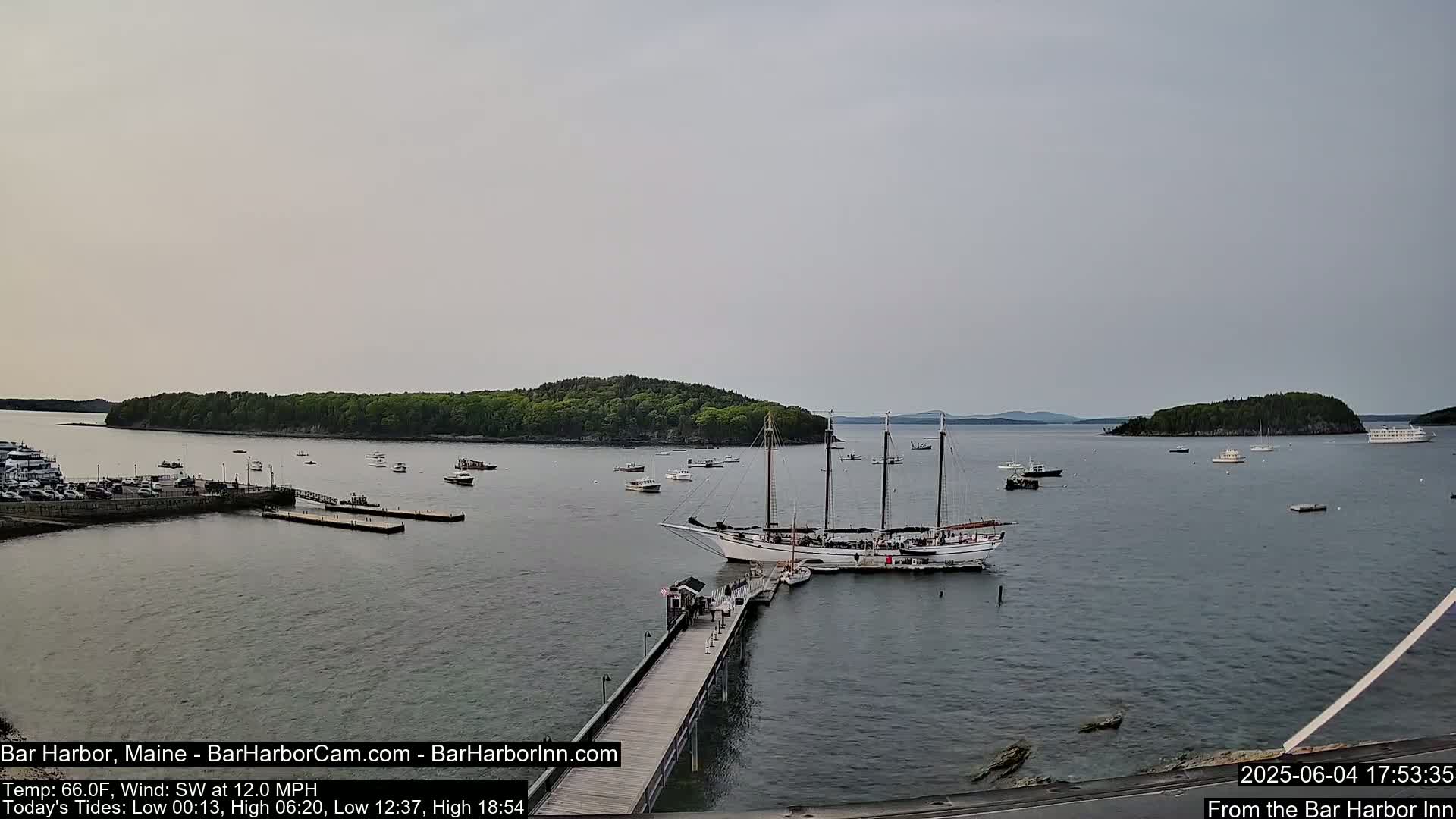 A large sailing ship is docked at a pier in a calm harbor, surrounded by smaller boats and islands under an overcast sky.