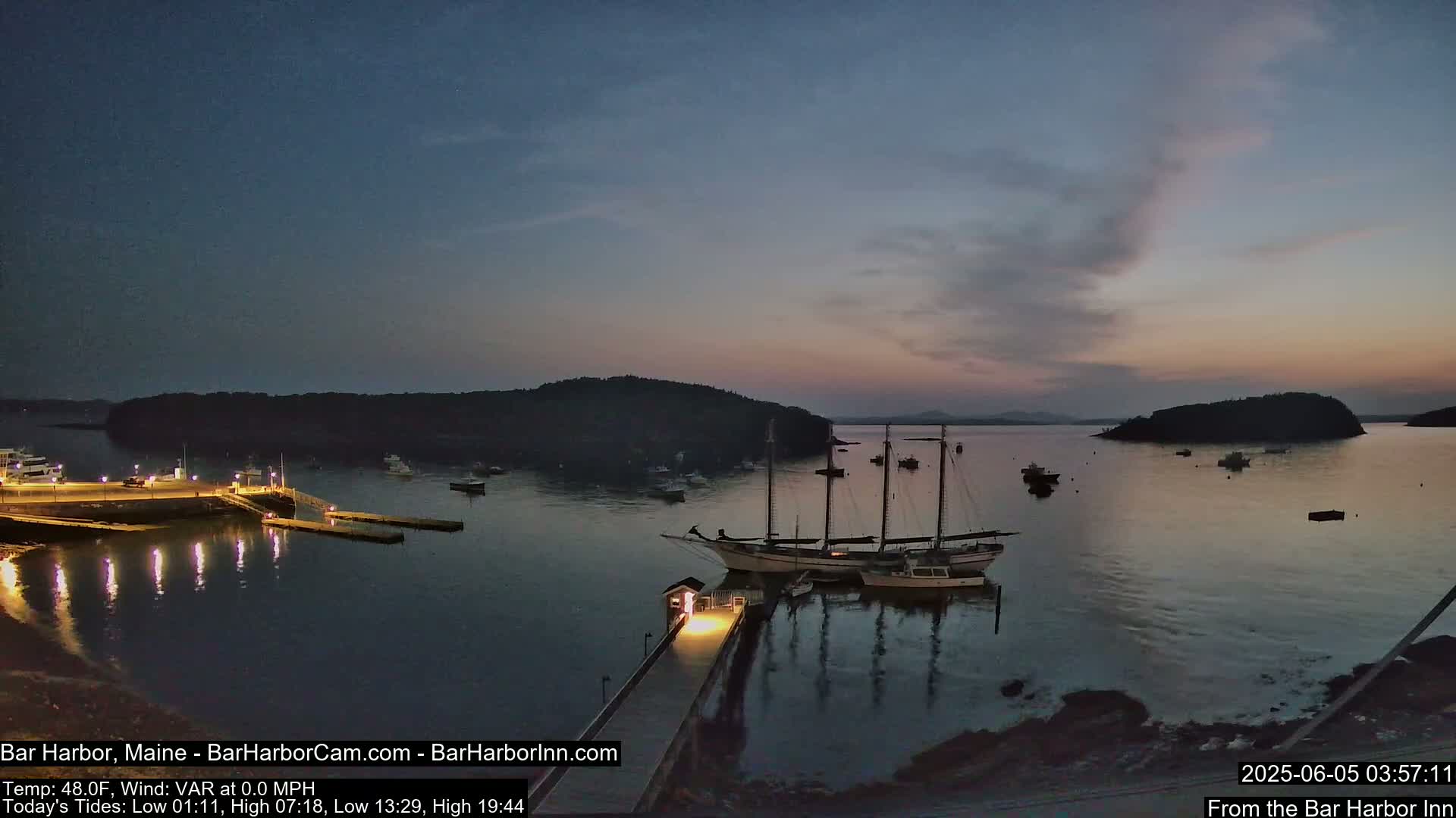 A calm harbor at twilight, with several sailboats and smaller vessels moored at a lit dock under a mostly clear sky.