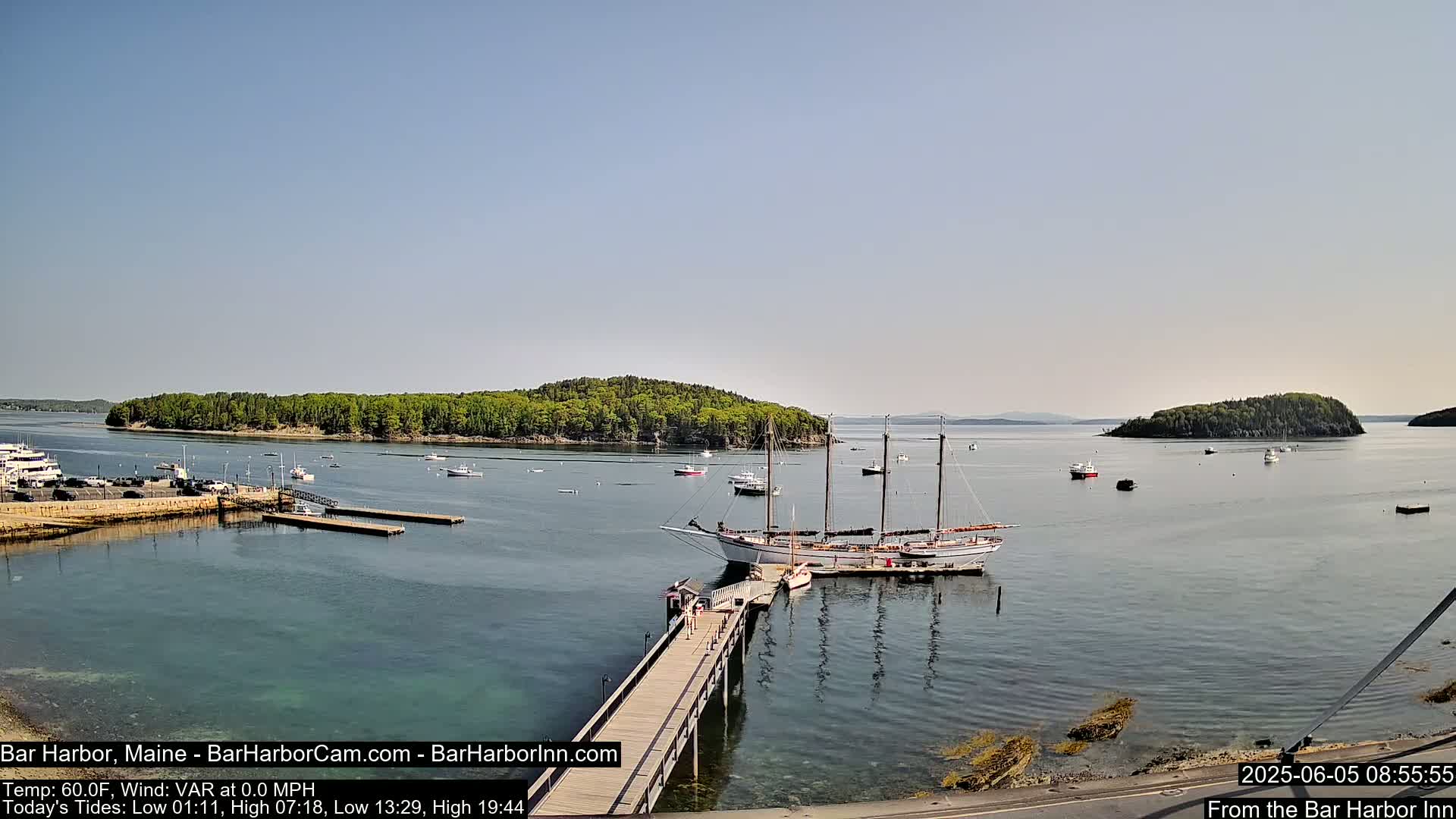 A calm harbor scene on a sunny day shows a large sailboat docked near a pier with several smaller boats and islands in the distance.