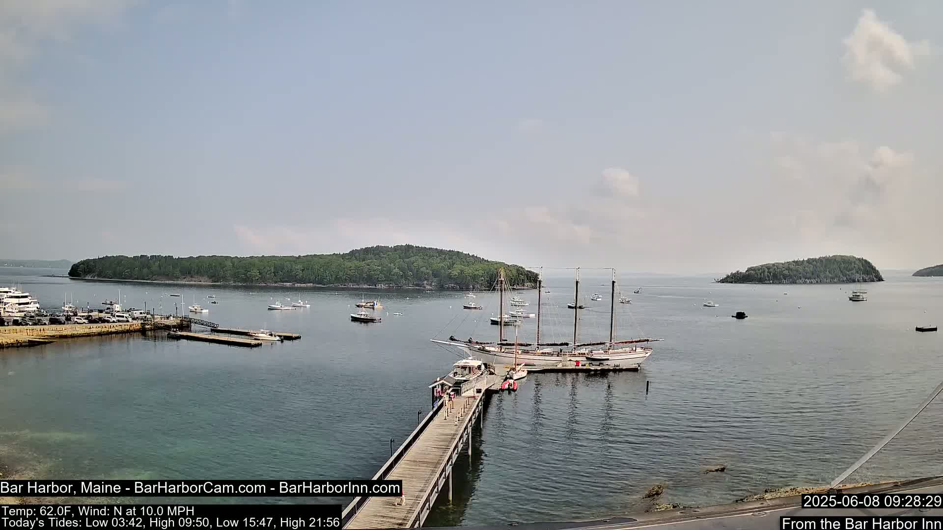 A calm, partly cloudy day shows a harbor with numerous boats, including a large sailing ship docked at a pier, and a small, tree-covered island in the distance.