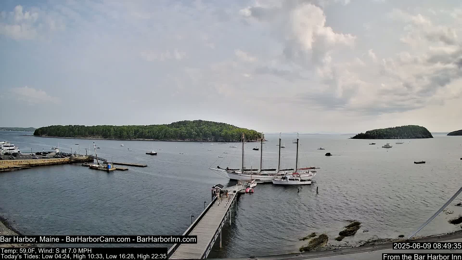A partly cloudy day shows a harbor with several boats, including a large sailing ship docked at a pier, with small islands visible in the distance.