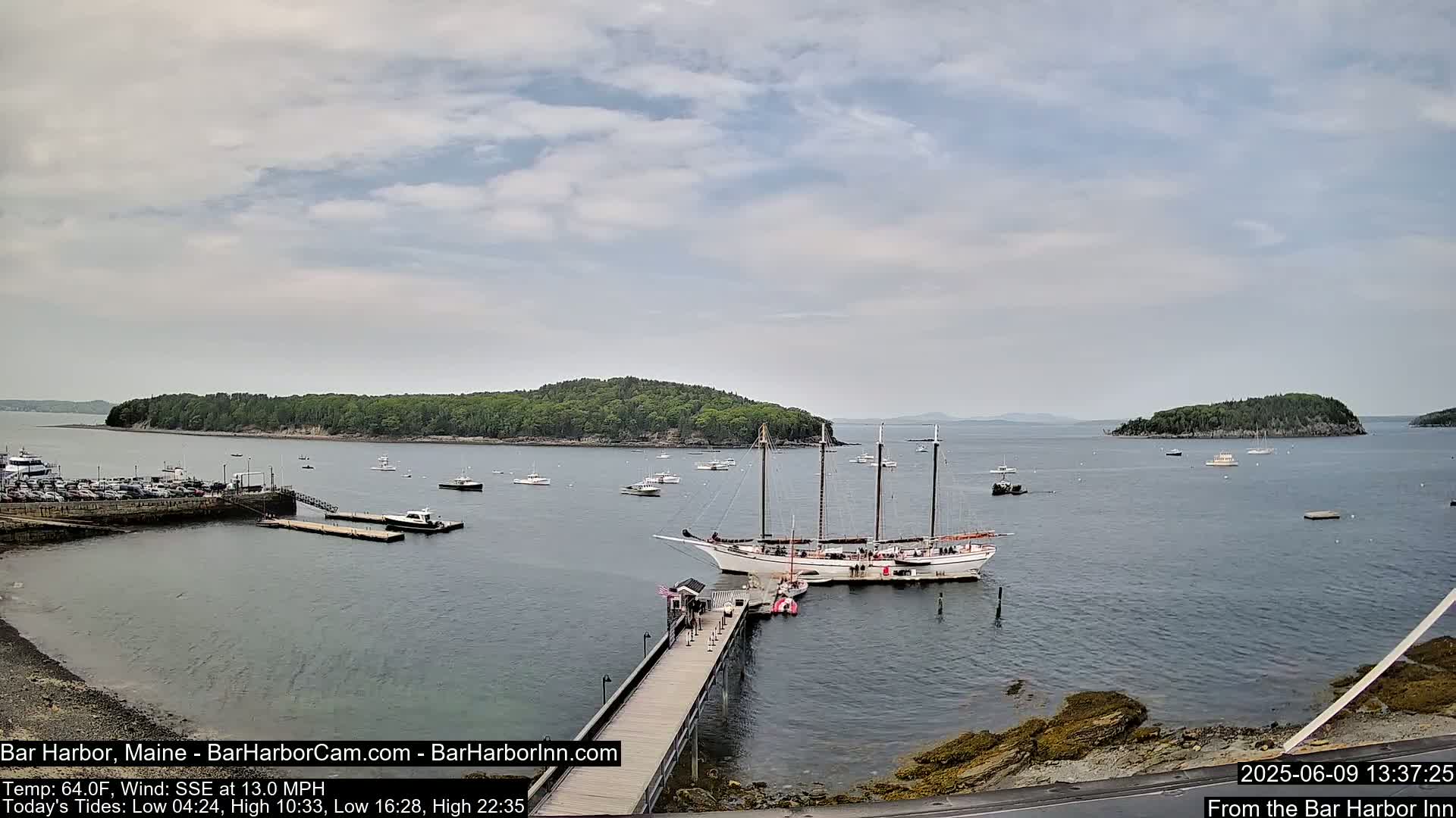 A large sailing ship is docked at a pier in a calm harbor, with several smaller boats and a wooded island visible in the distance under a mostly cloudy sky.