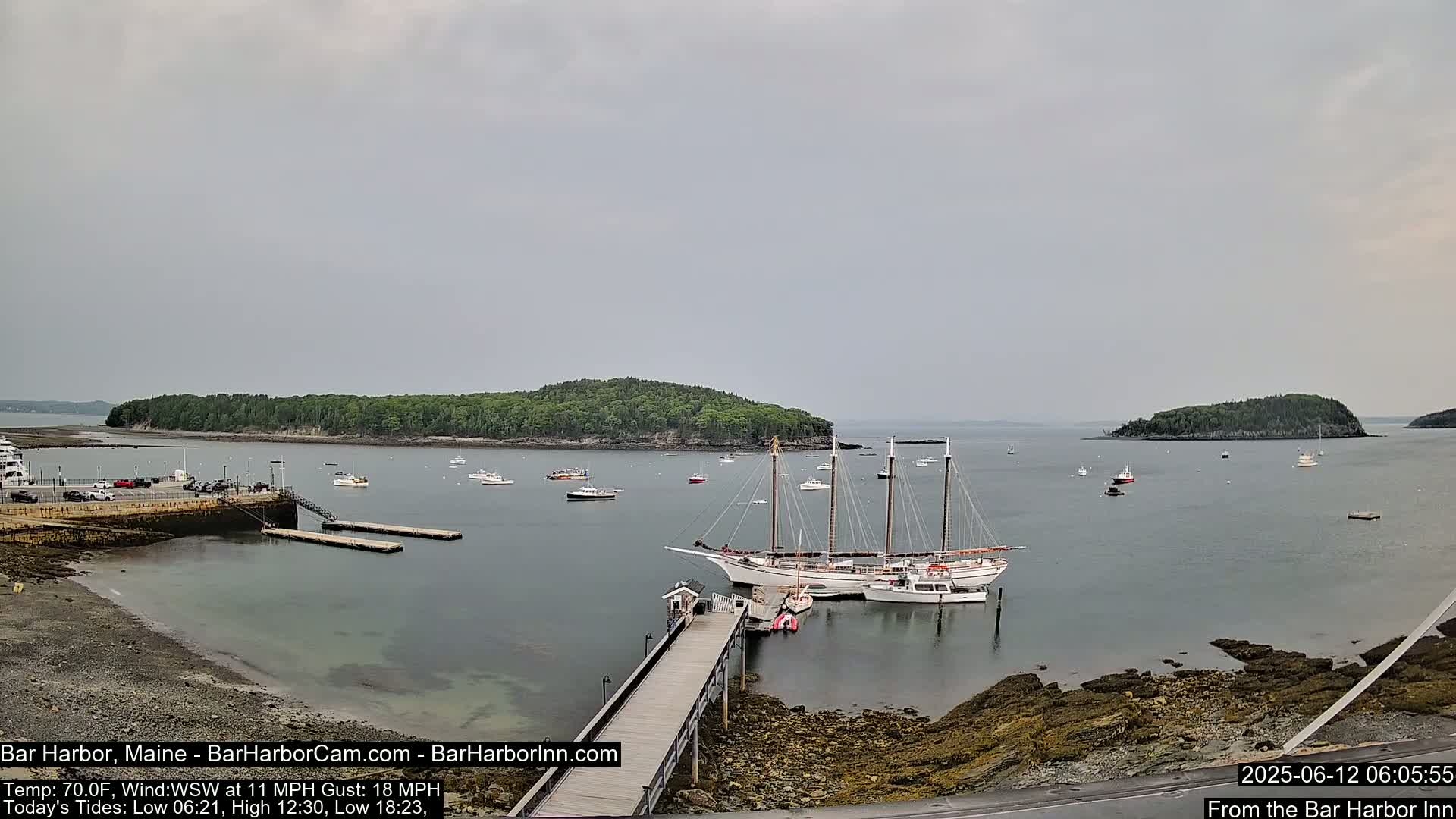 A large sailboat is docked at a pier near a rocky shoreline, with several smaller boats scattered in the calm, overcast harbor, surrounded by green islands.