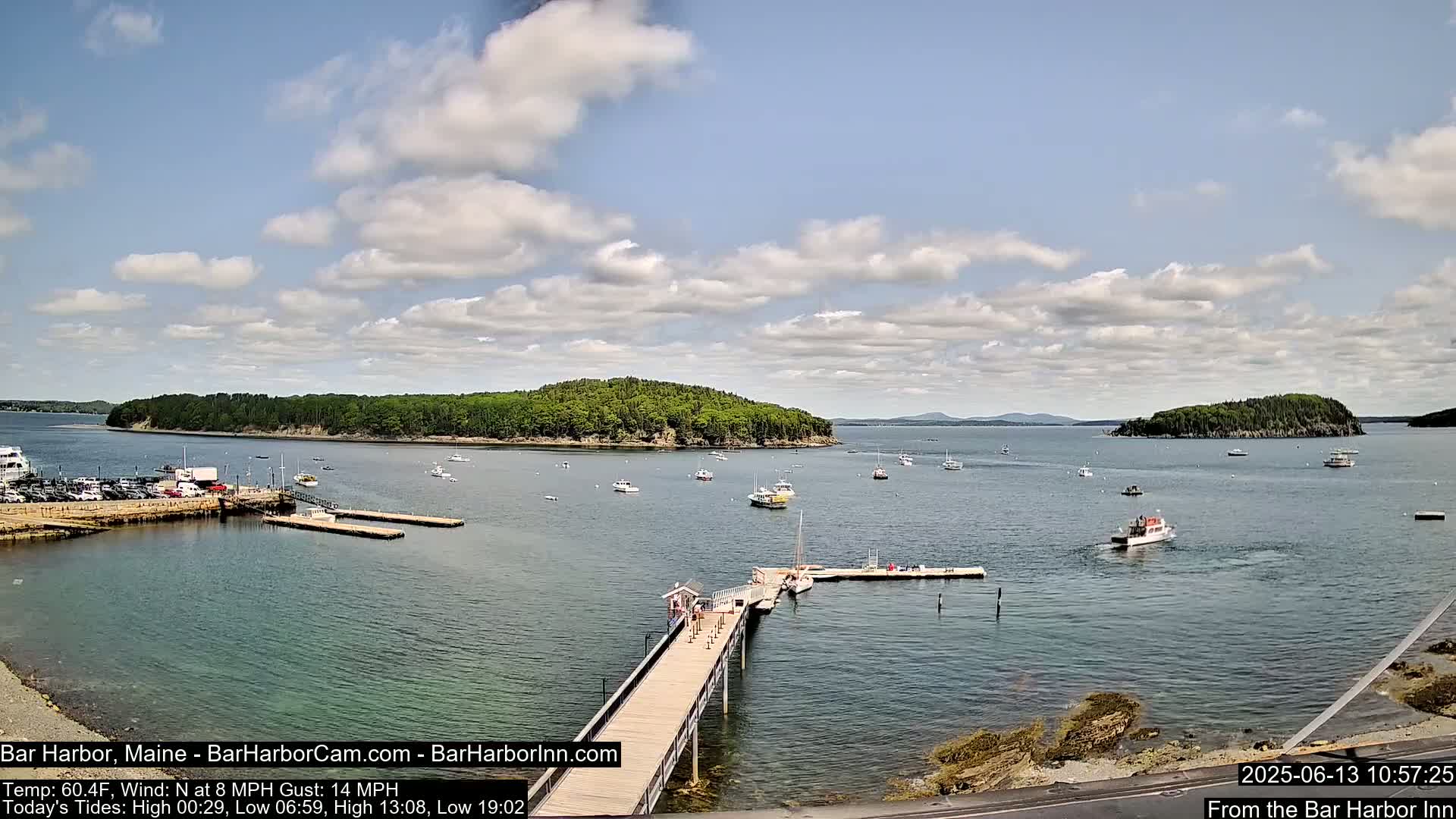 A partly cloudy day shows a harbor with several boats and a long wooden pier extending into the water, with islands and a distant mountain range visible in the background.