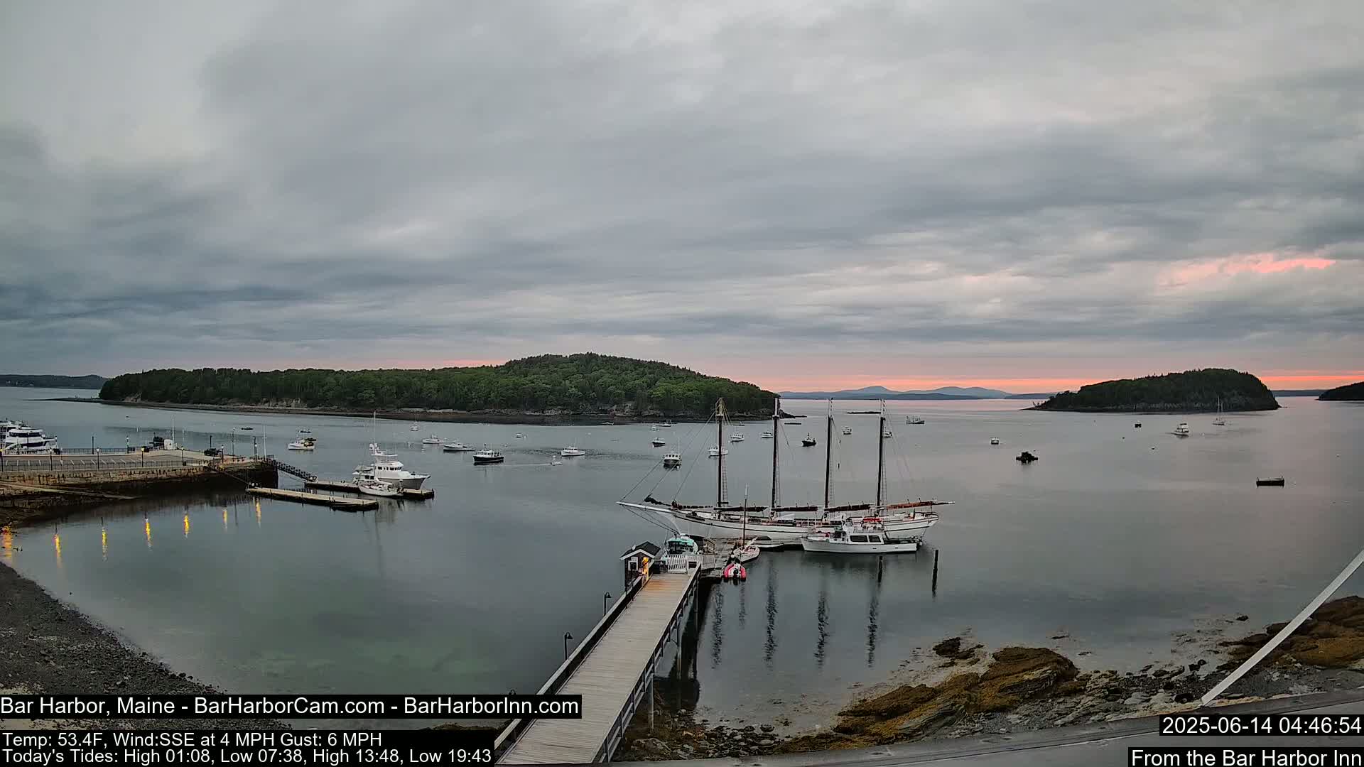 A calm harbor with numerous boats, including a large sailing ship, is seen at sunrise under a mostly cloudy sky.