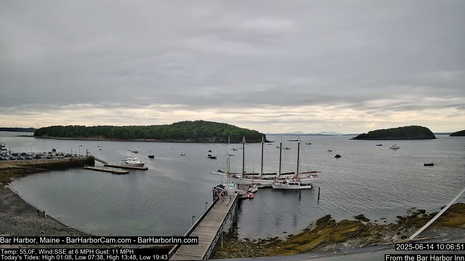 A calm, overcast day shows a harbor with several boats docked at a pier, including a large sailing ship, with islands and a distant mountain range visible across the water.