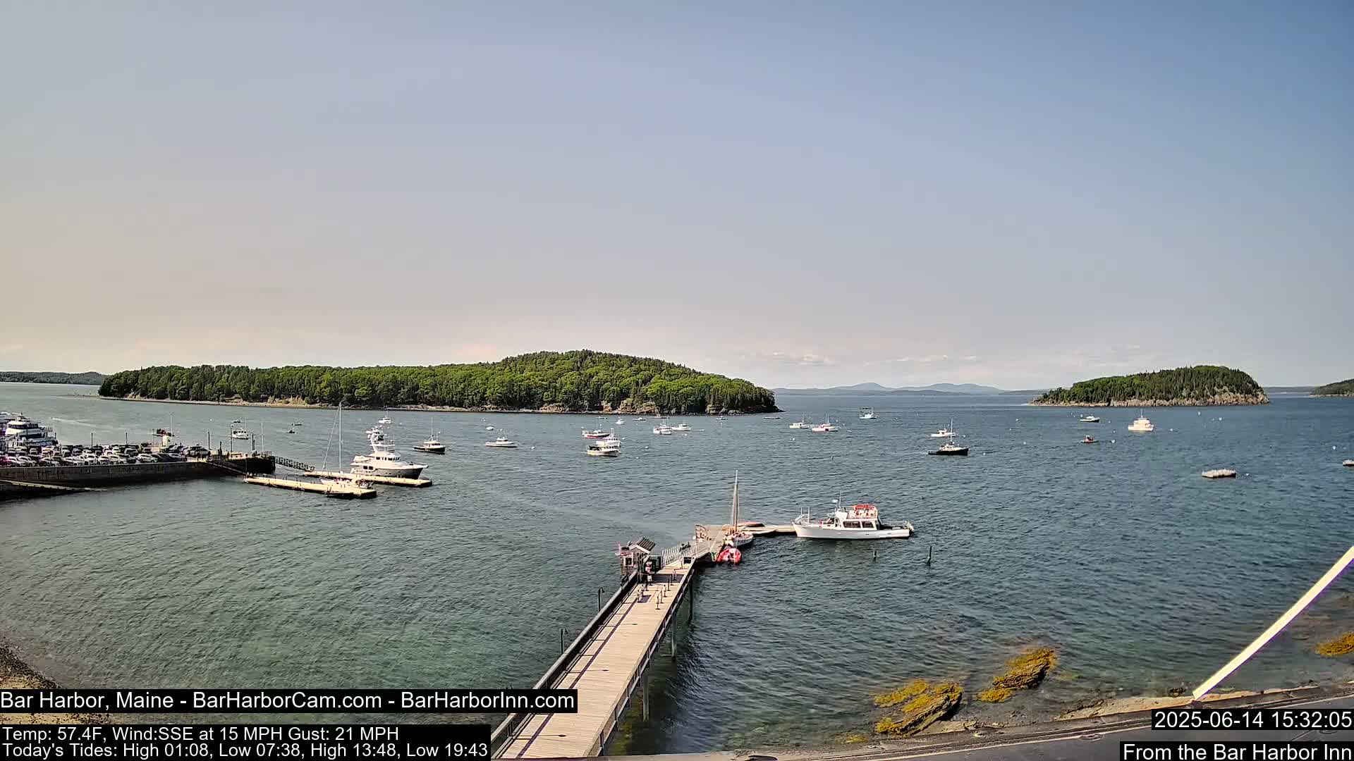 A calm, overcast day shows a harbor with several boats docked at a pier, including a large sailing ship, with islands and a distant mountain range visible across the water.