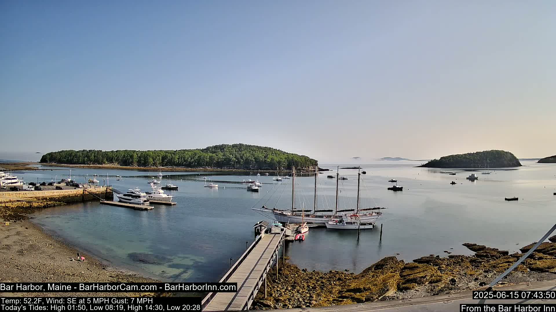 A calm harbor with numerous boats, including a large sailboat docked at a pier, is surrounded by rocky shores and verdant islands under a clear sky.