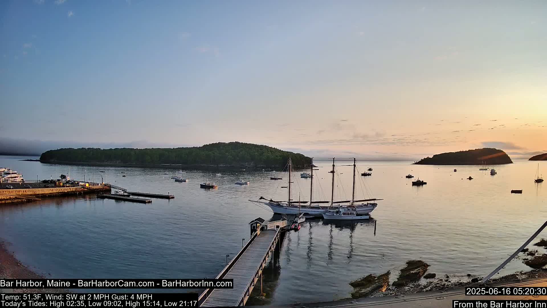 A calm harbor at sunrise, with several sailboats and motorboats moored near a wooden pier and a small, tree-covered island in the distance.
