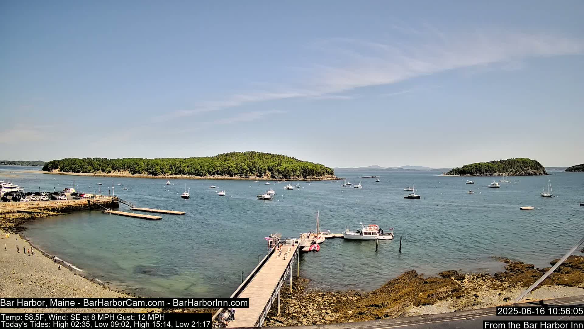 A sunny day reveals a harbor filled with boats near a small island, a wooden pier extending from a rocky shoreline, and a few people on the beach.