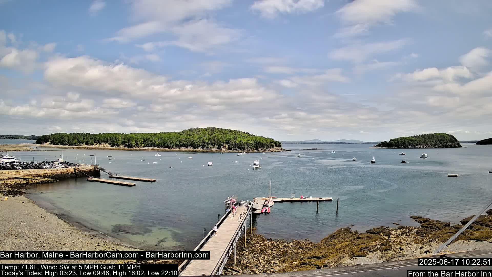 A partly cloudy day shows a harbor with boats and a long pier leading to a small building, with islands and a coastline in the background.