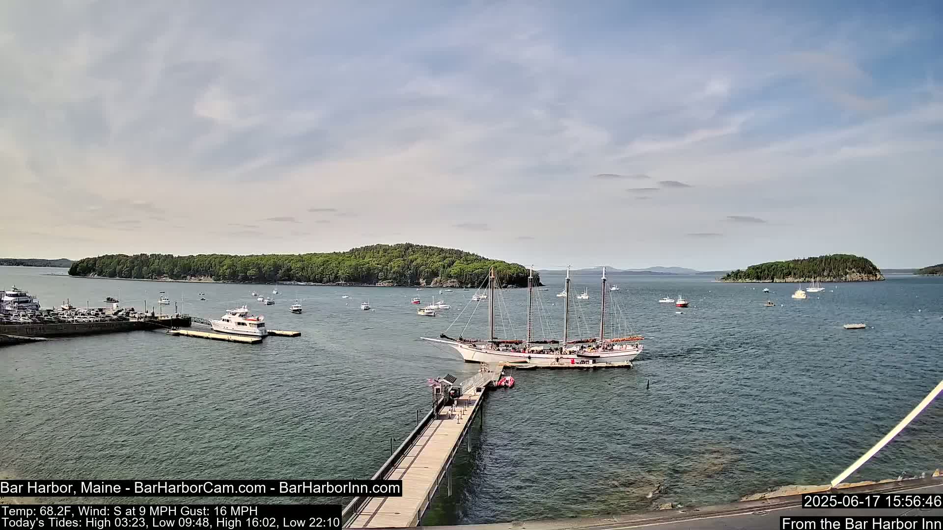 A large sailing ship is docked at a pier in a harbor with numerous smaller boats and islands in the background under a partly cloudy sky.