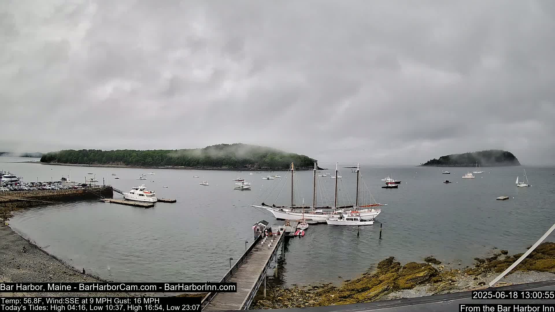 A large sailing ship is docked at a pier in a harbor on an overcast day, with several smaller boats and islands in the distance partially shrouded in fog.