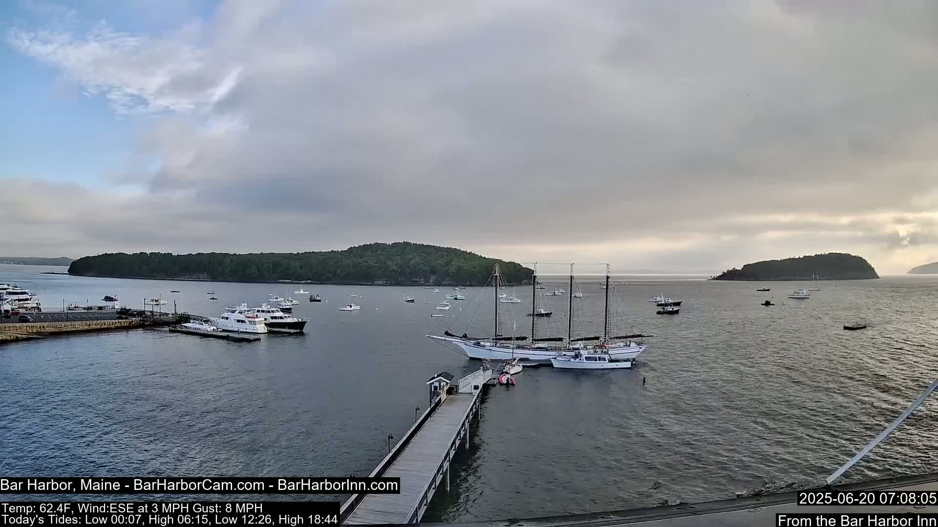 A calm harbor with numerous boats, including a large sailing ship moored at a pier, under a mostly cloudy sky.