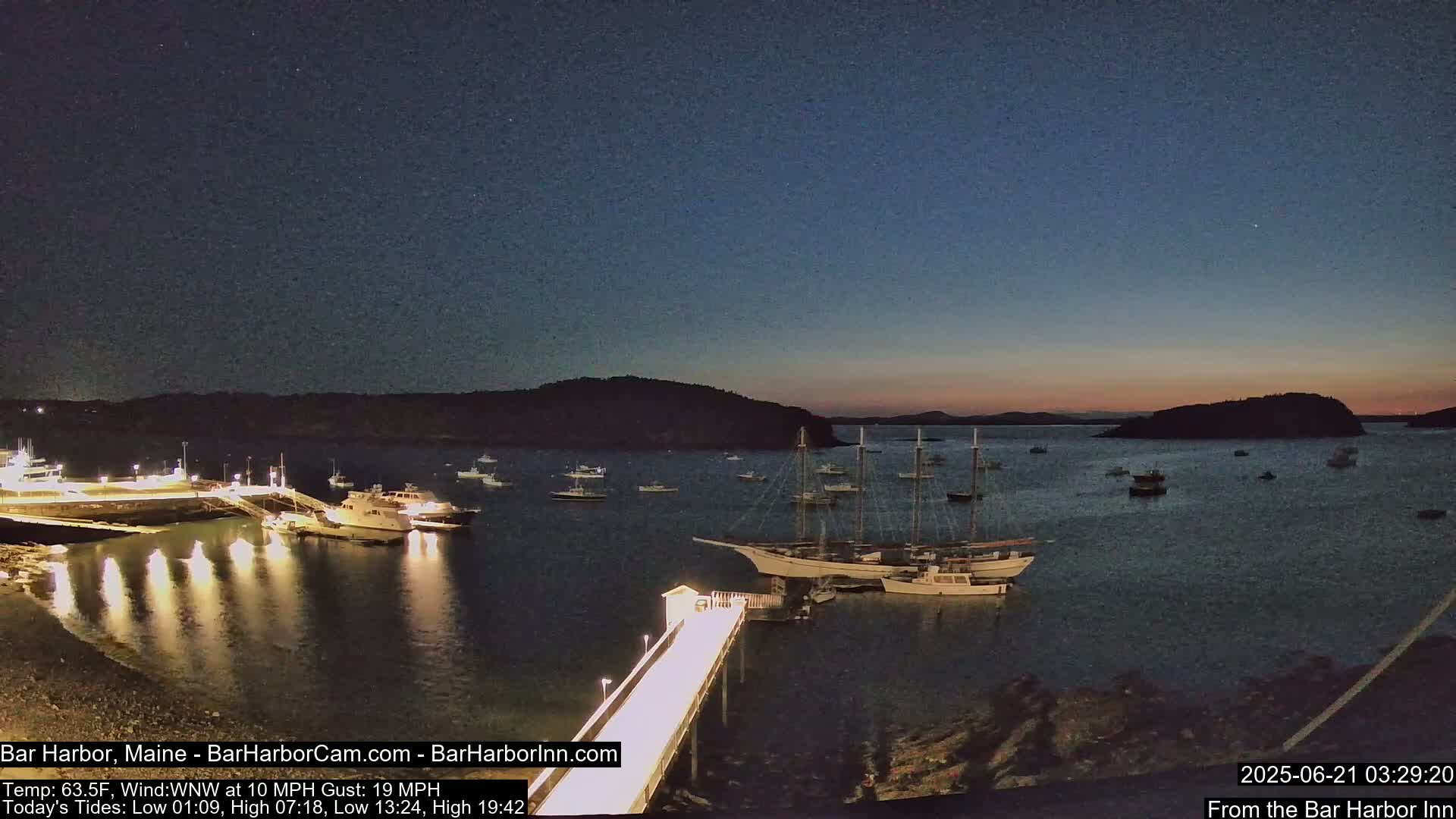 A brightly lit pier extends into a calm harbor at twilight, where several boats and a large sailing ship are moored near a shoreline with dark landmasses in the background.
