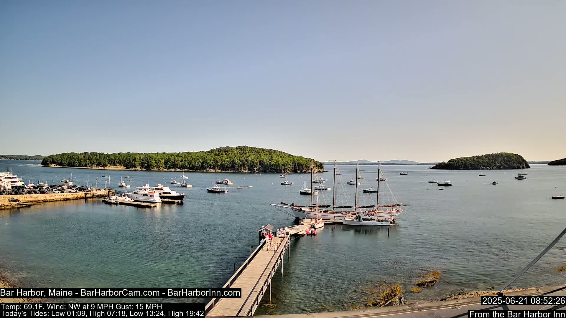 A calm harbor with numerous boats, including a large sailboat docked at a pier, is surrounded by islands under a clear blue sky.