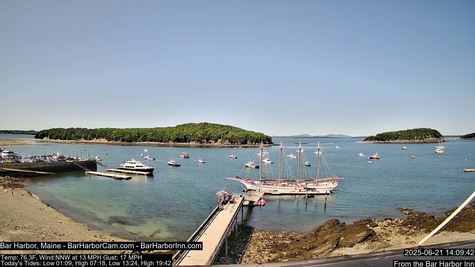 A harbor scene on a sunny day shows a large sailing ship docked at a pier, surrounded by smaller boats and a wooded island in the background.