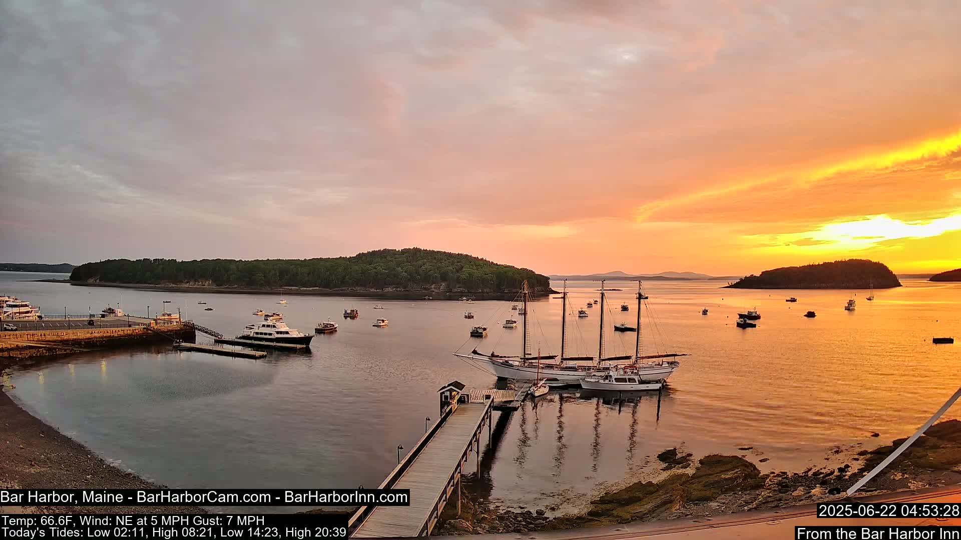 A calm harbor at sunset with several sailboats and motorboats moored at a pier, under a partly cloudy, orange-hued sky.